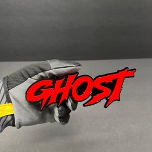 Könnte beinhalten: Ein rotes "GHOST"-Emblem mit schwarzem Umriss. Das Wort ist in einer fetten, stilisierten Schriftart gehalten. Das Emblem wird von einer behandschuhten Hand vor grauem Hintergrund gehalten.