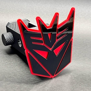 Puede incluir: Una cubierta para enganche de remolque en negro y rojo con el logotipo de Decepticon de Transformers. El emblema tiene una cara negra brillante con un borde rojo vibrante, montado sobre una base cuadrada negra texturizada con una perilla lateral.