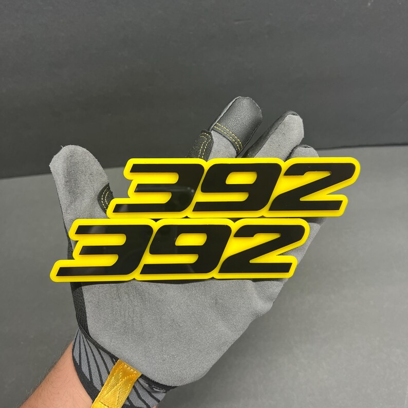 May include: Two yellow and black "392" emblems. The emblems are rectangular with rounded corners. The numbers are black and outlined in yellow. The emblems are held by a grey work glove.