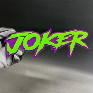 Peut inclure: Une enseigne superposée affichant le mot JOKER dans une police dentelée et stylisée. La couche supérieure est d'un vert citron vif, tandis que la couche inférieure est d'un violet profond, créant un contraste marqué aux bords anguleux.