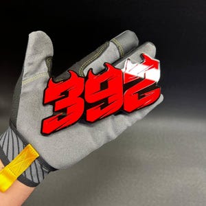 May include: A red and white "392" emblem with a flame design, held in a gray work glove. The emblem has a glossy finish and is set against a dark background. The glove has yellow accents.