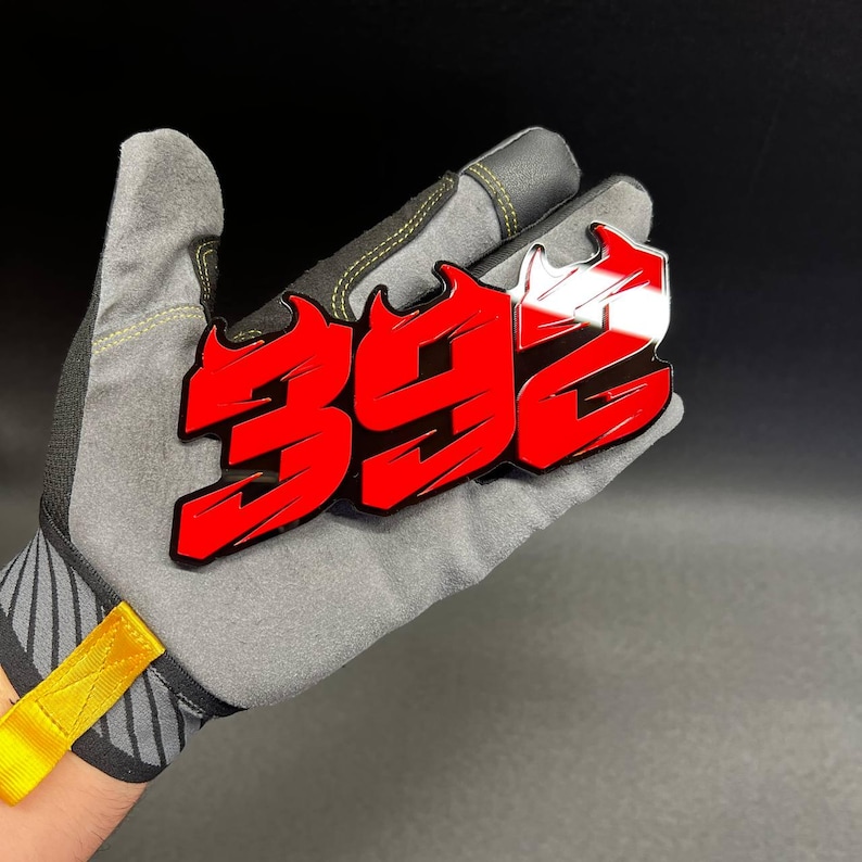May include: A red and white "392" emblem with a flame design, held in a gray work glove. The emblem has a glossy finish and is set against a dark background. The glove has yellow accents.