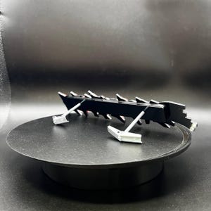 May include: A black, angular object with a series of sharp, triangular projections along its top edge. The object is supported by two silver-coloured, angled supports. The item is displayed on a black, circular platform.