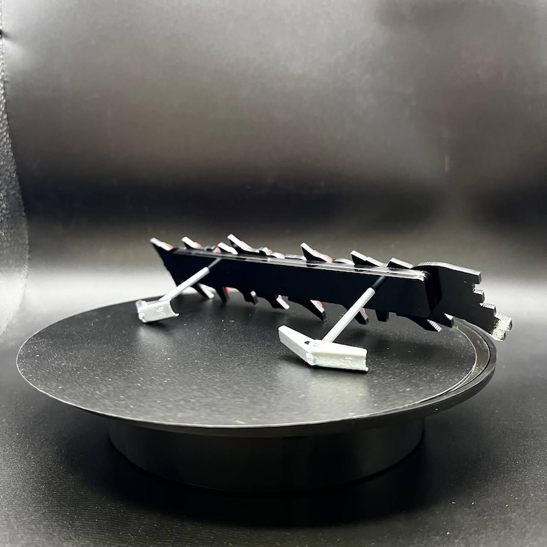May include: A black, angular object with a series of sharp, triangular projections along its top edge. The object is supported by two silver-coloured, angled supports. The item is displayed on a black, circular platform.