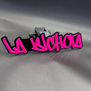 以下が含まれることがあります： 鮮やかなピンクのグラフィティ風の文字で「LA BICHOTA」と書かれた黒いヒッチカバー。デザインは黒い長方形のベースに取り付けられており、背面には車両に取り付けるための質感のある円筒形のノブが付いています。