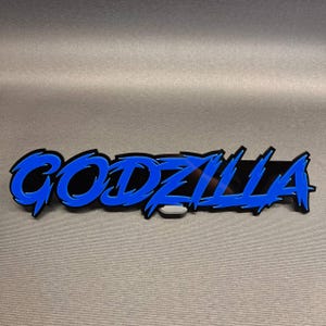 Op de afbeelding: Een op maat gemaakt bord met het woord GODZILLA in een grillig, gestileerd lettertype. De letters bestaan uit een felblauwe toplaag met scherpe, puntige randen, gemonteerd op een zwarte achtergrond die de contouren van de tekst volgt.