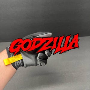 May include: A red "GODZILLA" logo with a black outline. The logo is held by a person wearing a black and gray work glove with a yellow strap.