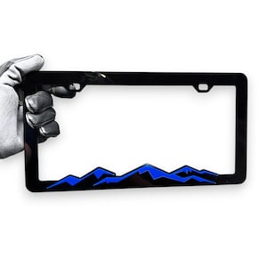 Può includere: Cornice nera per targa con un design a catena montuosa blu. La cornice è rettangolare con finitura lucida. La catena montuosa blu è nella parte inferiore.