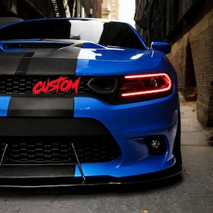 Puede incluir: Un coche deportivo azul con franjas negras y la palabra "CUSTOM" en rojo en la parrilla delantera. El coche tiene luces LED rojas y un parachoques delantero negro. La imagen está tomada en un callejón.