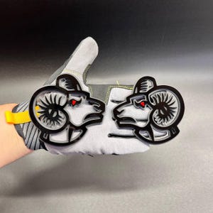 May include: Two layered acrylic emblems featuring a stylised ram head design. Each emblem consists of a black outer frame, a light grey middle layer, and a small red accent for the eye. The ram head is depicted in profile with curved horns.