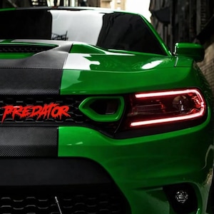 Puede incluir: Primer plano de un coche verde con una franja negra en el centro. La palabra "PREDATOR" está en rojo en la parte delantera. Los faros del coche emiten un brillo rojo.
