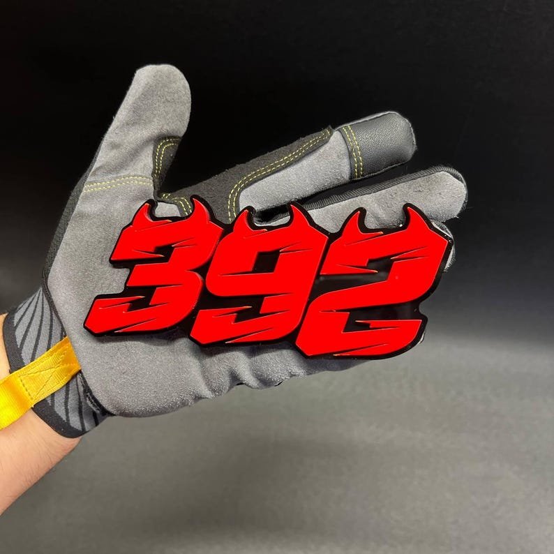 May include: A red and black emblem with the number "392" in a bold, stylized font. The emblem is held in a gray work glove with black accents and yellow stitching. The background is dark.