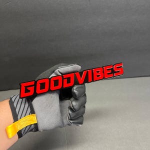 May include: A black and red "GOOD VIBES" emblem is held by a gloved hand. The text is in a bold, sans-serif font with a red outline and a black interior. The glove is gray and black with a yellow strap.