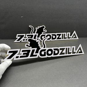 May include: Two white and black decals with the text "7.3L GODZILLA" and a Godzilla silhouette. The decals are rectangular with a stylized font and a black Godzilla graphic. The background is a solid gray.