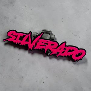 以下が含まれることがあります： 黒い縁取りが施された鮮やかなピンク色の滴るようなフォントで「SILVERADO」という文字がデザインされた金属製エンブレム。エンブレムはダークグレーの取り付けブラケットに固定されており、側面には固定用の質感のある円筒形のノブが2つ付いています。