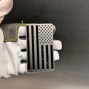 May include: A rectangular black metal plate featuring a cutout design of the United States flag. The design includes vertical stripes and a field of stars, all rendered in a solid matte black finish with clean, precise edges throughout the piece.