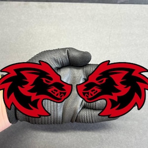 May include: Two red and black dragon head decals. The dragon heads are facing each other. The decals are held by a black glove. The background is a light gray color.