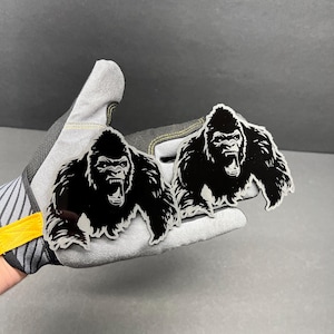 May include: Two black and white gorilla-shaped decals. The gorilla is depicted with an open mouth and a fierce expression. The decals are held in a gray work glove.