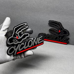May include: Black and silver Cyclone logo patch featuring a cartoon rabbit with red eyes and the word "Cyclone" in a stylized font with a red underline. The patch is held by a gloved hand.