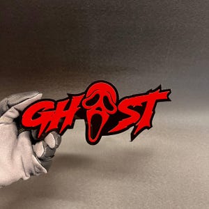 May include: A red and black acrylic sign featuring the word GHOST in a jagged, stylised font. The letter O is replaced by a red and black mask graphic with elongated eye and mouth openings, creating a sharp, high-contrast visual design.
