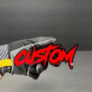 May include: A red "CUSTOM" sticker with a dripping effect, held by a gray and black glove. The sticker has a black outline and is against a dark gray background. The glove has yellow accents.