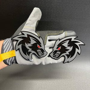 Puede incluir: Dos emblemas idénticos de cabeza de lobo con un diseño en capas de color negro brillante y gris, rasgos estilizados afilados y ojos rojos penetrantes. Las cabezas de lobo están colocadas una al lado de la otra, mostrando una estética audaz.