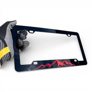 May include: Black license plate frame with a red mountain range design. The frame is rectangular with four mounting holes and a glossy finish. The mountain range is a stylized, three-dimensional design.