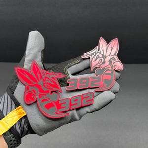 May include: Two red and gray stylized hornet emblems with the number "392" are displayed. The emblems are held in a gray work glove. The hornet design is a bold, graphic representation.