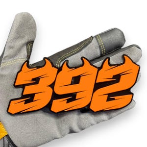 May include: An orange and black emblem with the number "392" is displayed. The emblem is set against a gray work glove. The numbers are in a bold, stylized font with a flame-like design.