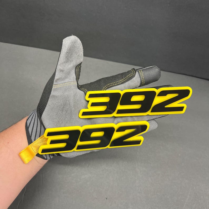 May include: Two black and yellow 392 emblems are held in a grey work glove. The emblems have a glossy finish and are in a sans-serif font. The glove has black accents and a yellow pull tab.
