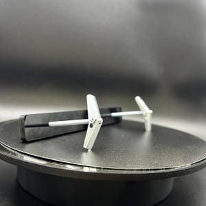 May include: Two silver drywall anchors with a butterfly design, each with a screw. One anchor is partially inserted into a black, rectangular object. The anchors are on a black surface.