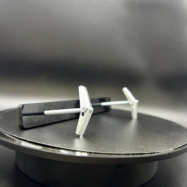 May include: Two silver drywall anchors with a butterfly design, each with a screw. One anchor is partially inserted into a black, rectangular object. The anchors are on a black surface.