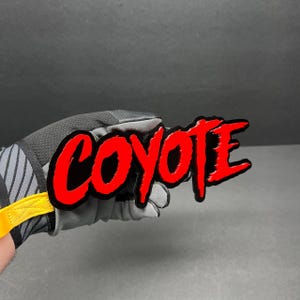 May include: A black and red "COYOTE" sticker with a bold, stylized font. The sticker is held by a gray and black glove with a yellow strap. The background is a solid, dark gray.