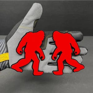 Puede incluir: Dos siluetas planas de color rojo brillante de una criatura tipo Bigfoot, cada una representada en perfil de caminata, están colocadas sobre un guante de trabajo gris y negro que presenta costuras amarillas y una lengüeta amarilla en la muñeca.