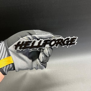 Può includere: Logo "HELLFORGE" in bianco e nero. Il logo è tenuto da un guanto grigio e nero con accenti gialli. Il logo è un marchio denominativo stilizzato con un carattere in grassetto e leggermente usurato.