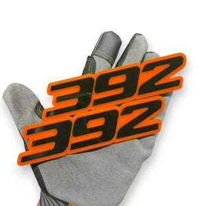 May include: Two orange and black "392" emblems. The emblems are in a bold, stylized font. The emblems are held by a grey work glove.