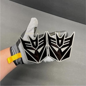 Könnte beinhalten: Zwei schwarze und silberne Decepticon-Logo-Embleme. Die Embleme werden in einem grauen Arbeitshandschuh mit gelbem Riemen gehalten. Das Decepticon-Logo ist ein stilisiertes, eckiges Design mit schwarzem Hintergrund und silbernen Akzenten.