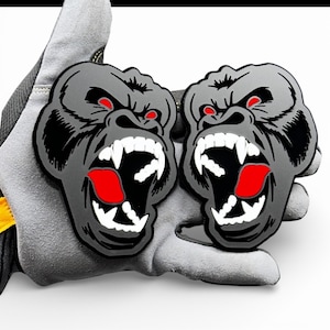 May include: Two grey gorilla-faced patches with red eyes and open mouths, revealing white teeth and red tongues. The patches are held by a grey work glove.