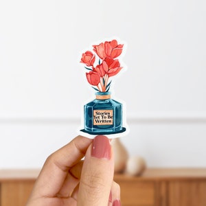 May include: A blue glass bottle with a label that reads "Stories Yet To Be Written" and a bouquet of red flowers inside. The bottle is on a white background.
