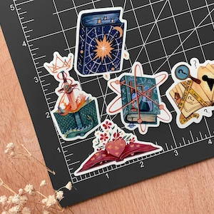 May include: Five illustrated book stickers with different themes. The stickers feature a book with a crown and a sword, a book with a heart, a book with a magnifying glass, a book with an atom, and a book with a star.