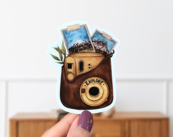 May include: A vintage-style camera with a brown leather case and the word "EXPLORE" printed on the front. Two photos are tucked into the top of the case, one showing a mountain range and the other showing a mountain peak.