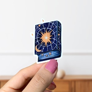 May include: A blue book sticker with a celestial design. The cover features a large, orange star with a white centre and a crescent moon in the bottom left corner. The background is a dark blue with white stars and lines.