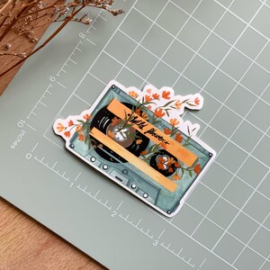 May include: A vintage cassette tape with a blue and white design, surrounded by orange and yellow flowers. The tape label reads "Wild Flowers".