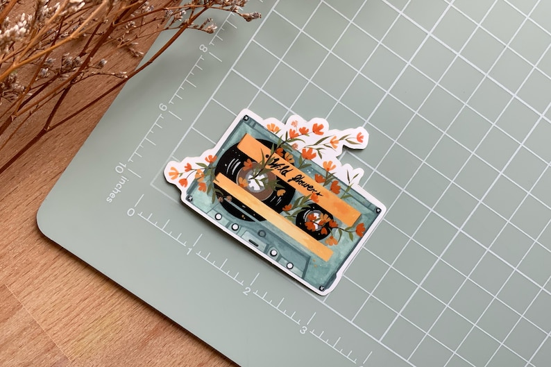 May include: A vintage cassette tape with a blue and white design, surrounded by orange and yellow flowers. The tape label reads "Wild Flowers".