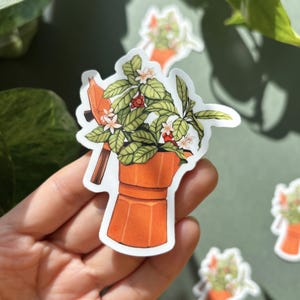Peut inclure: Un autocollant orange repr&eacute;sentant une cafeti&egrave;re avec une plante qui en sort. La plante a des feuilles vertes, des fleurs blanches et des baies rouges. D'autres autocollants avec des motifs similaires sont visibles.