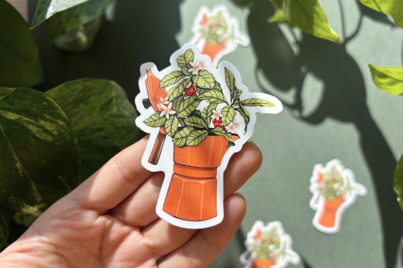 Peut inclure: Un autocollant orange repr&eacute;sentant une cafeti&egrave;re avec une plante qui en sort. La plante a des feuilles vertes, des fleurs blanches et des baies rouges. D'autres autocollants avec des motifs similaires sont visibles.