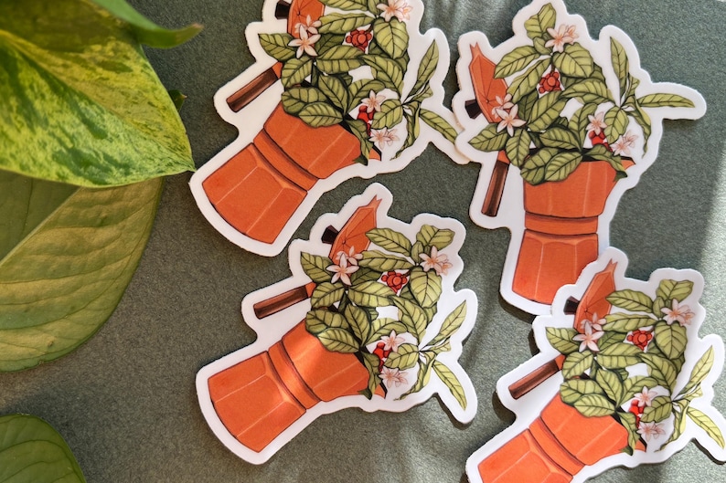 Peut inclure: Quatre autocollants repr&eacute;sentant une cafeti&egrave;re orange avec un arrangement floral. Les autocollants ont une bordure blanche et sont dispos&eacute;s sur une surface verte. L'arrangement floral comprend des feuilles vertes, des fleurs blanches et des baies rouges.
