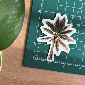 May include: A palm tree sticker with a white border, placed on a green cutting mat. The sticker is approximately 2 inches tall. A green plant leaf is visible in the upper left corner, and the background is a wooden surface.
