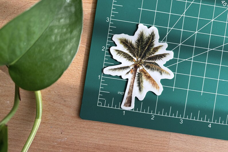 May include: A palm tree sticker with a white border, placed on a green cutting mat. The sticker is approximately 2 inches tall. A green plant leaf is visible in the upper left corner, and the background is a wooden surface.