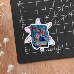 May include: A sticker of a closed book with a red atomic symbol around it. The book has a blue cover with handwritten notes and drawings of scientific equipment, including a beaker, a flask, and a graduated cylinder. The book is titled "The Book of Science".